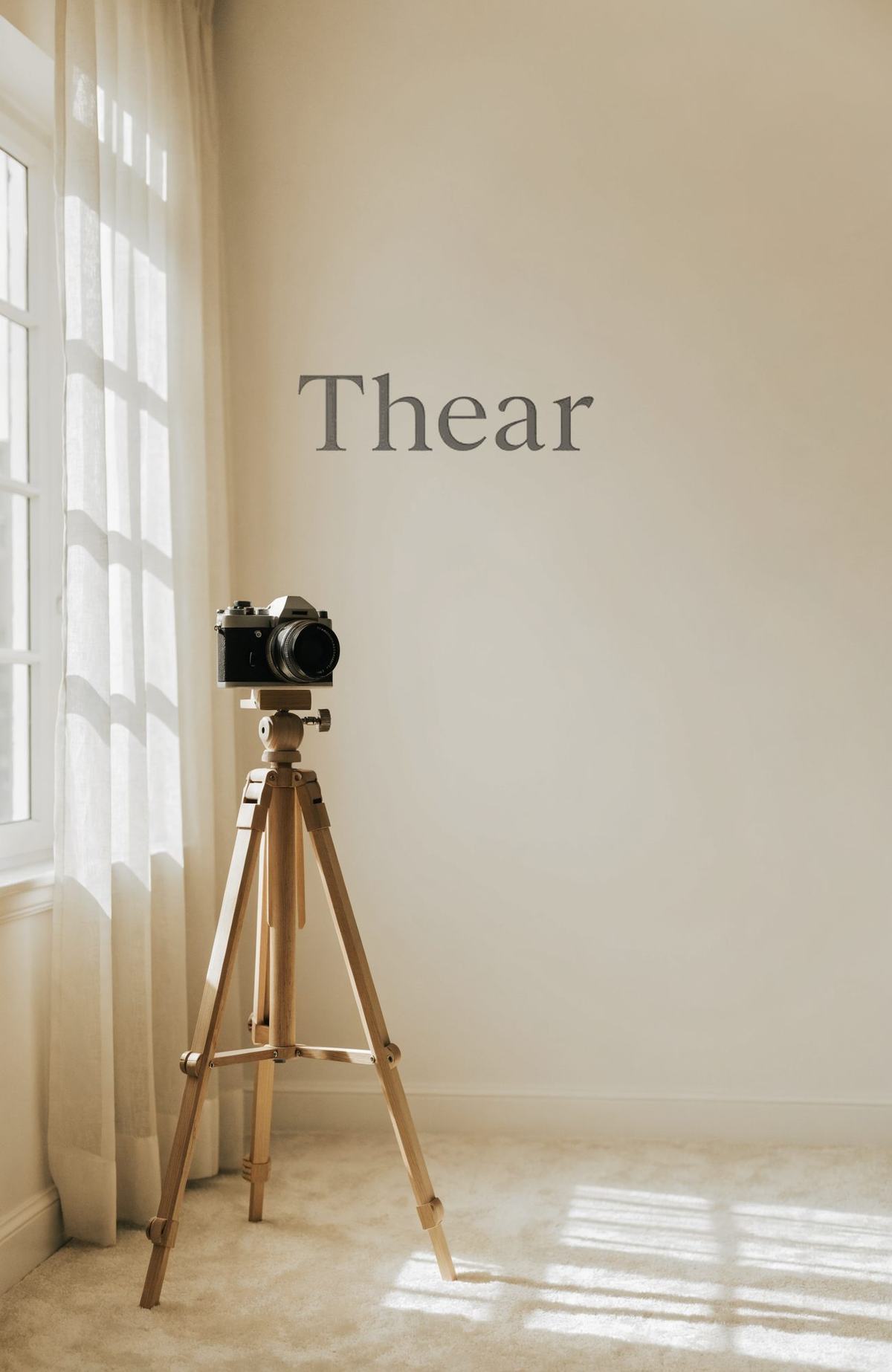 아이보리 커튼이 드리운 창가에 세워 둔 카메라 삼각대 — 벽 가운데에 Thear 워드마크