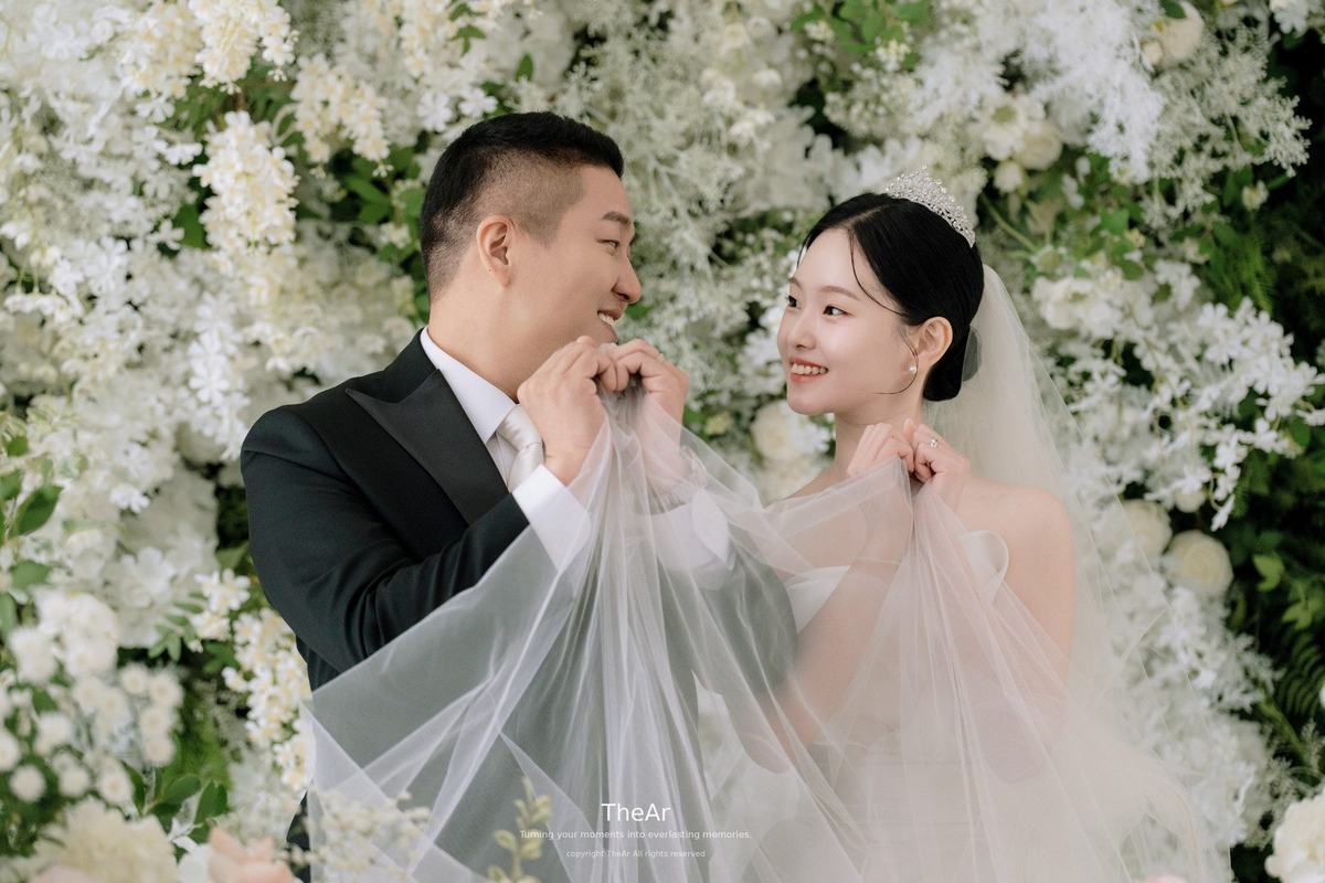 흰 꽃벽 앞에서 신부의 베일을 들어 올리며 마주 보는 신랑