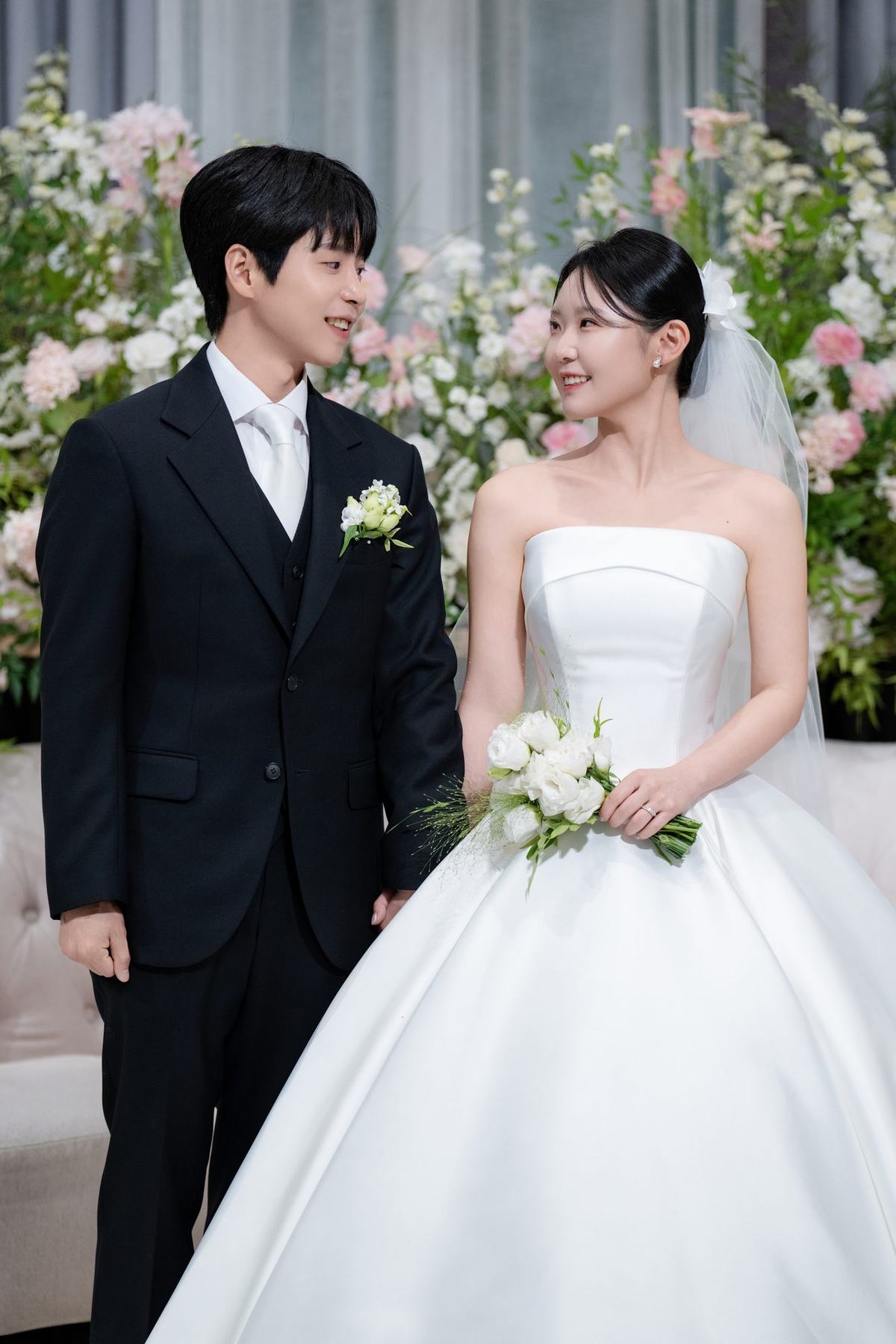 분홍빛 생화를 배경으로 부케를 든 신부와 나란히 서서 마주 보는 신랑