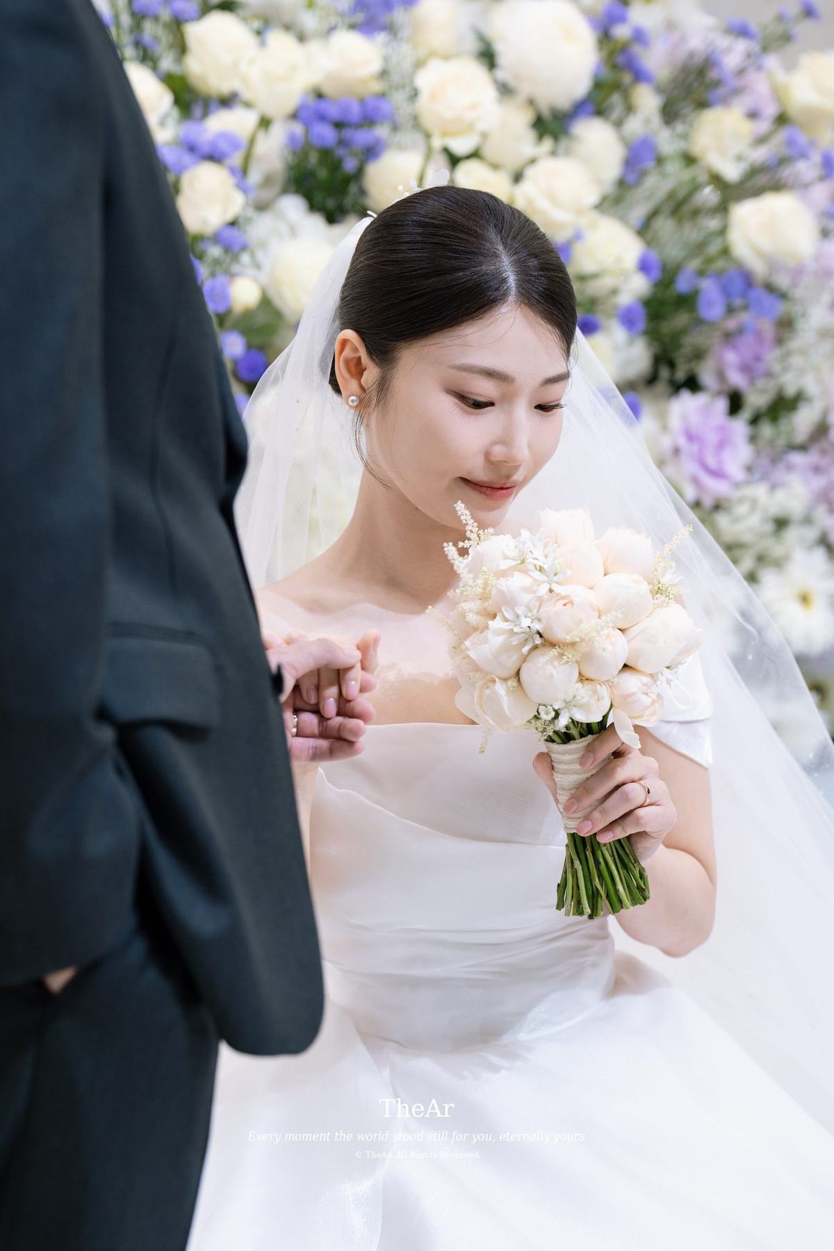 신랑의 어깨 너머로 보이는, 흰 꽃 부케를 든 신부의 손과 내려다보는 눈