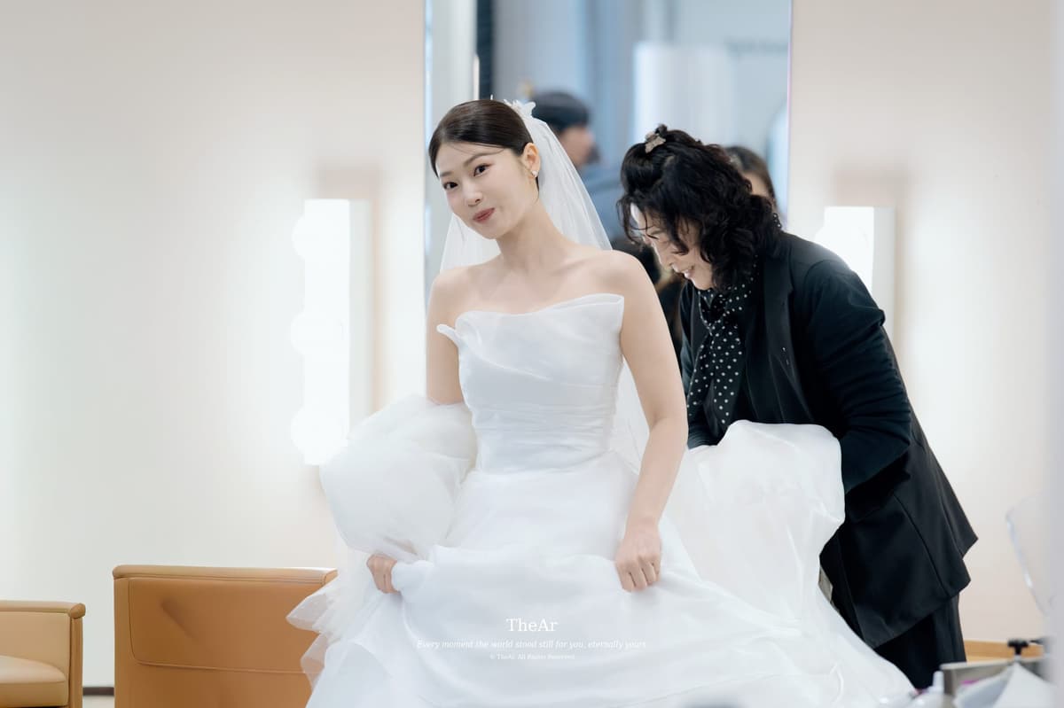 메이크업샵 창가에서 드레스 자락을 모아 매만져 주는 손과 그 앞에 선 신부