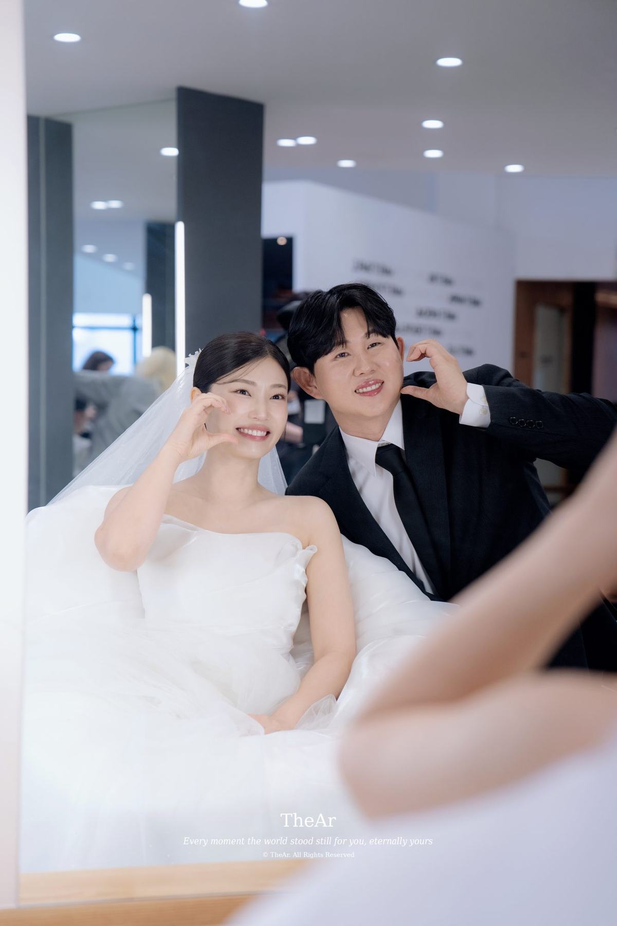 메이크업샵 거울 앞에 앉은 신부가 손하트를 만들고 그 뒤에서 신랑이 따라 웃는다
