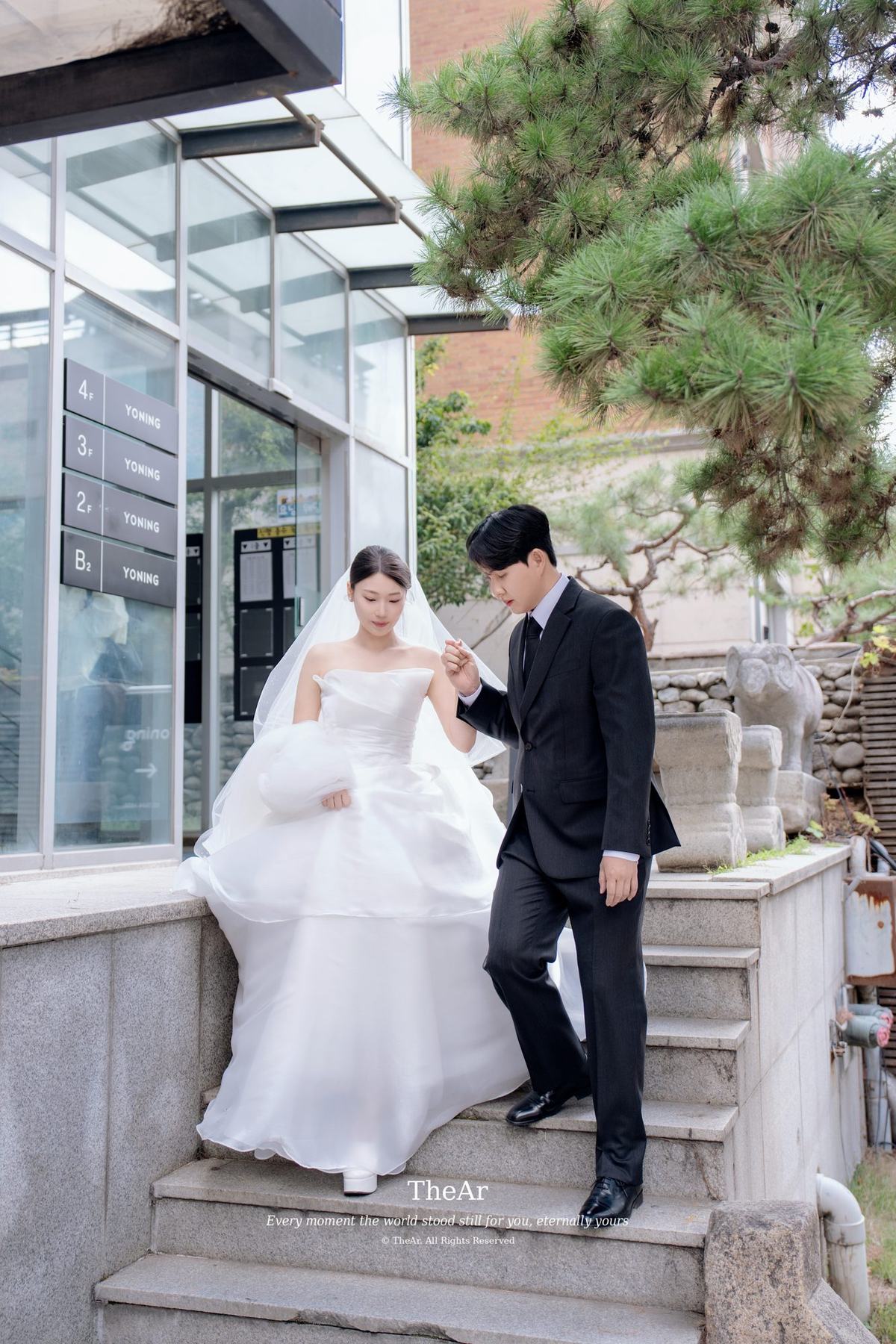 유리 지붕이 덮인 건물 입구 계단을 신랑의 손을 잡고 내려오는 신부