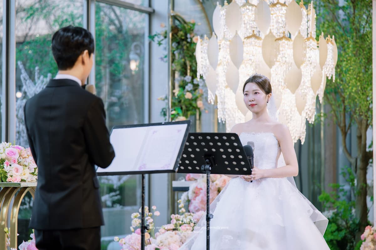 L65 가든홀 샹들리에 아래 보면대 앞에서 혼인서약서를 읽는 신랑과 마이크를 들고 마주 선 신부