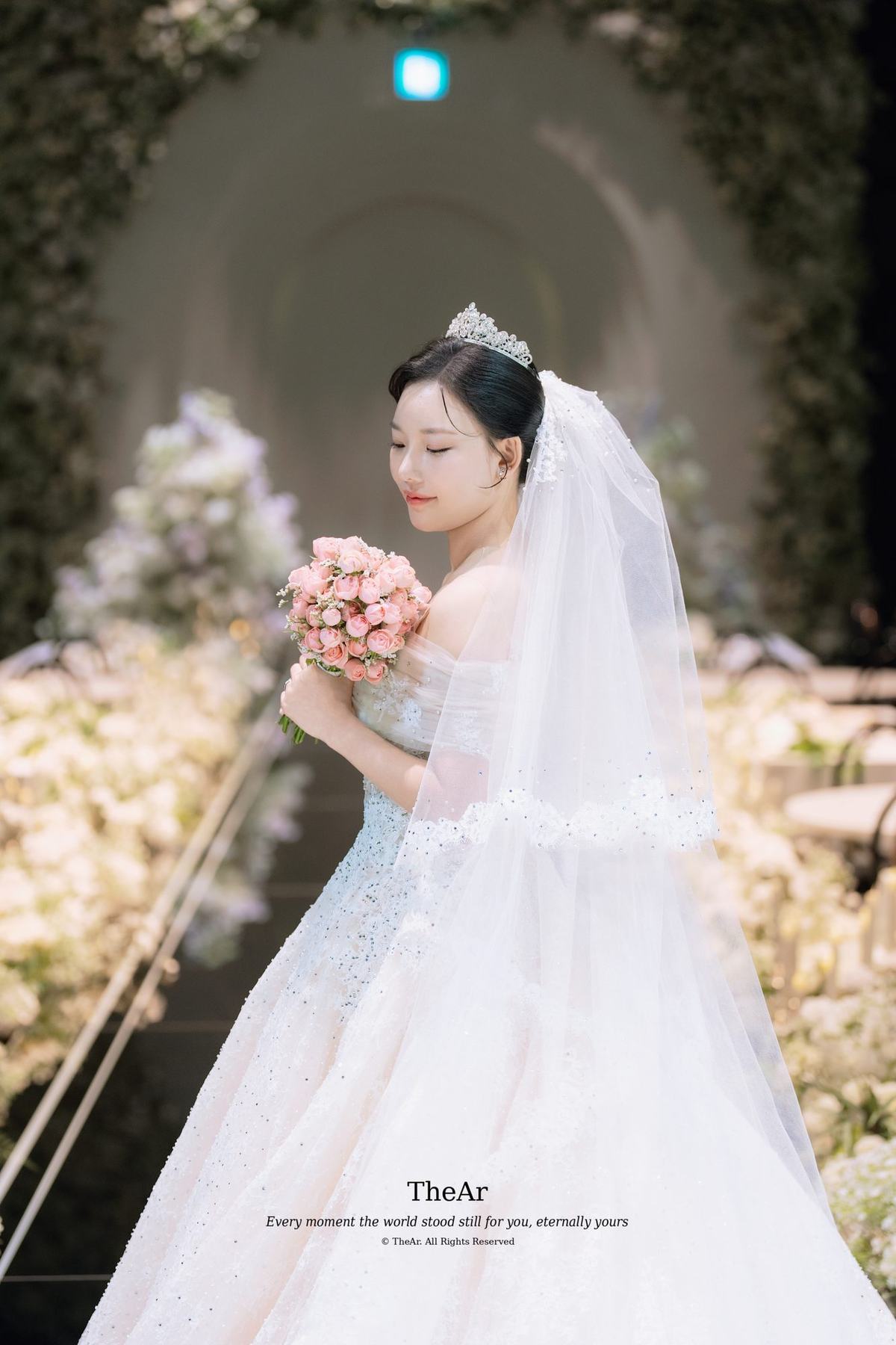 꽃으로 덮인 아치 아래에 혼자 서서 부케를 품에 안은 신부