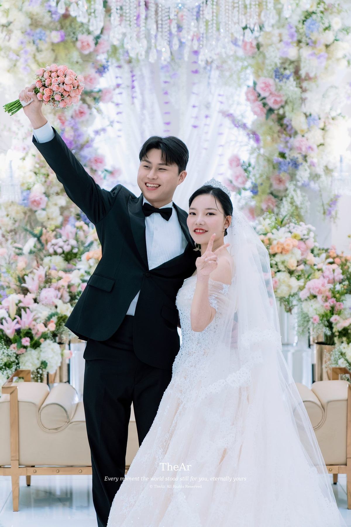 신부대기실 꽃벽 앞에서 부케를 높이 들어 올린 신랑과 그 옆에서 웃는 신부