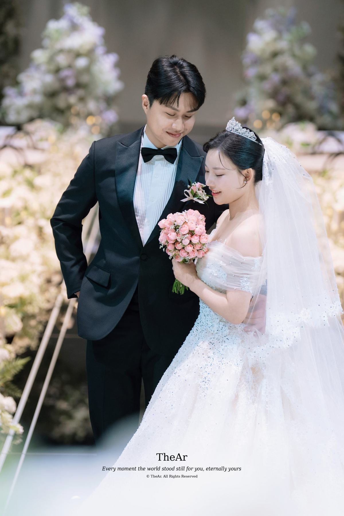 꽃 장식을 배경으로 서로에게 몸을 기울여 눈을 맞추는 신랑과 부케를 든 신부