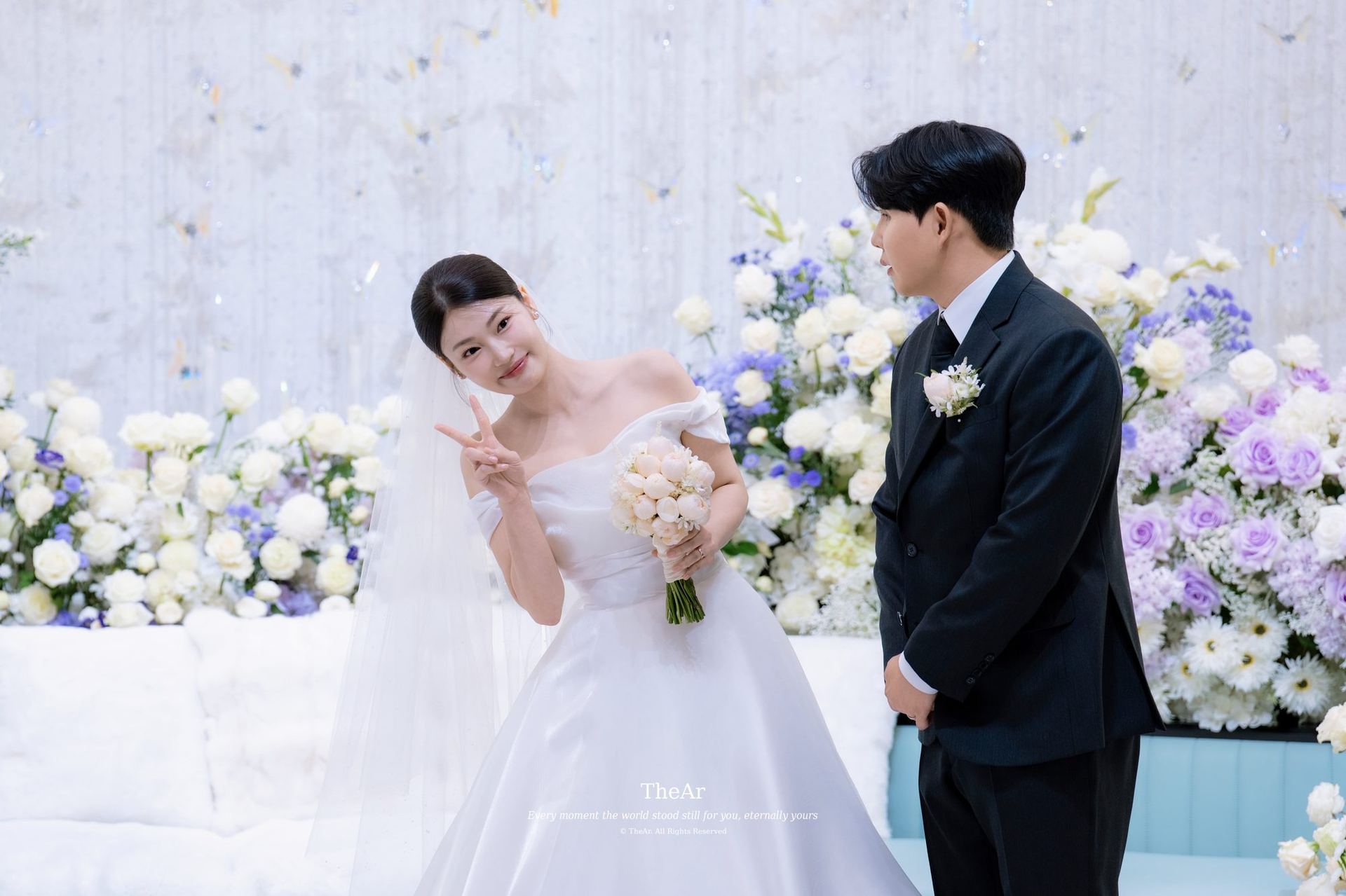 꽃으로 가득한 L65 컨벤션홀 대기실 벽 앞에서 부케를 든 신부와 그를 바라보며 웃는 신랑